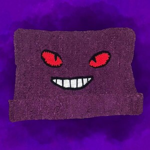 May include: A purple beanie featuring a stitched design of a cartoon face with red eyes and a wide, toothy grin. The hat is made of a textured material and is set against a purple background.