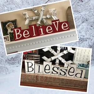 Op de afbeelding: Twee decoratieve houten borden met de woorden "Believe" en "Blessed" in witte letters. Het "Believe" bord heeft rode blokken, terwijl het "Blessed" bord witte blokken heeft. Een grote houten sneeuwvlok en een kalender zijn ook zichtbaar.