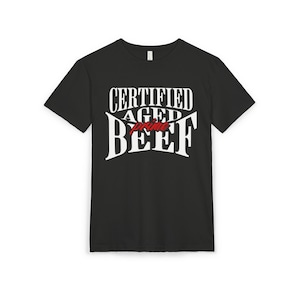 May include: Black t-shirt featuring the text "CERTIFIED AGED prime BEEF" in white and red. The design uses a bold, vintage font. The t-shirt has a crew neck and is made of a soft material.
