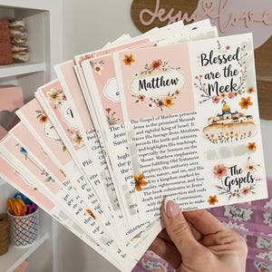 Könnte beinhalten: Ein Stapel rosa und weißer Karten mit floralen Designs und Text. Die Karten zeigen das Wort "Matthew" und den Satz "Blessed are the Meek". Die Karten werden in einer Hand gehalten.