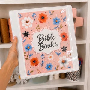 May include: A white three-ring binder featuring a pink cover with a floral pattern of red, blue, and white flowers. The center of the cover displays a decorative white label with a scalloped border containing the black text Bible Binder.