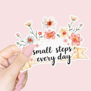 May include: A floral sticker with the text "small steps every day" in black script. The sticker features a banner design with watercolour flowers in shades of pink, orange, and white. The background is a soft pink.