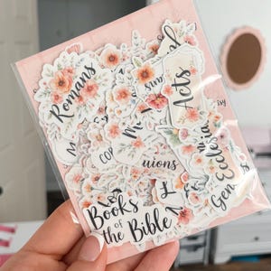 May include: A package of floral stickers featuring the names of Bible books. The stickers have a white background with watercolor-style flowers in shades of peach, pink, and green. The text is in a handwritten style font.