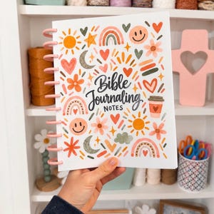 May include: A white spiral-bound notebook with pink plastic rings featuring the text Bible Journaling NOTES in black script. The cover is decorated with colorful illustrations of suns, rainbows, hearts, flowers, smiley faces, and coffee cups.