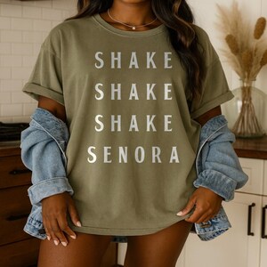 May include: Olive green t-shirt with the words "SHAKE SHAKE SHAKE SENORA" printed in silver. The shirt has short sleeves and a relaxed fit. A light blue denim jacket is draped over the shoulders.