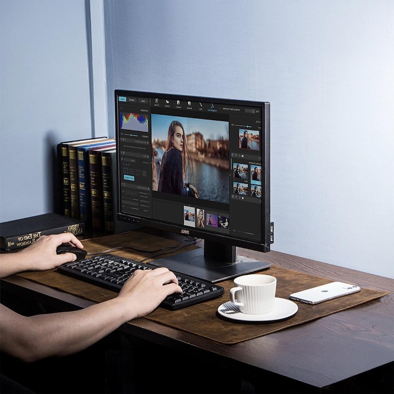 May include: A computer monitor displaying photo editing software sits on a wooden desk. A black keyboard, mouse, and a white coffee cup are on the desk. A brown leather desk mat and a stack of books are also visible.
