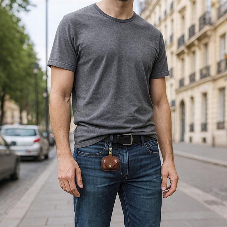 May include: A brown leather case with a gold-colored clasp is attached to a black belt. The case is small and round, with a button closure. The person is wearing a gray t-shirt and blue jeans.