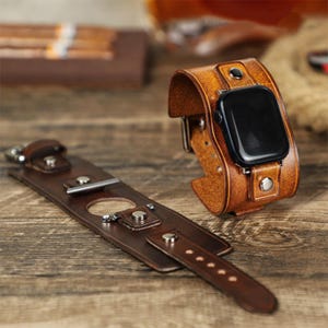 May include: A brown leather Apple Watch band with a black watch face. The band is a cuff style with a buckle closure and metal rivets. A separate, matching leather piece with a buckle and metal hardware is also present.