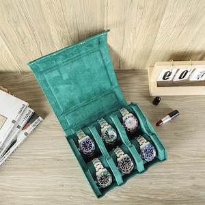 May include: A teal green watch case with six silver wristwatches with various colored watch faces. The case is open, revealing the watches nestled in individual compartments. A wooden calendar and lipstick are in the background.