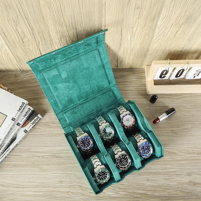 May include: A teal green watch case with six silver wristwatches with various colored watch faces. The case is open, revealing the watches nestled in individual compartments. A wooden calendar and lipstick are in the background.