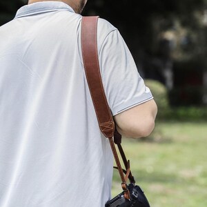 May include: A brown leather camera strap with a stitched design is draped over a person's shoulder. The strap is attached to a black camera. The person is wearing a light blue polo shirt.