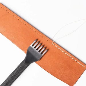 May include: A tan leather strip with a straight line of white stitching along the edge, positioned next to a black metal leather stitching chisel with six prongs and a textured handle, accompanied by a thin white thread loop.