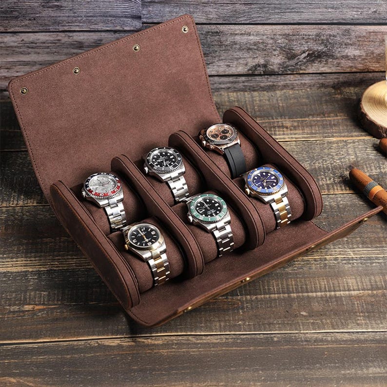 May include: A brown, open watch case displaying six wristwatches with silver and gold bands. The watches have various face colors, including black, blue, and green. The case is made of a soft, suede-like material.