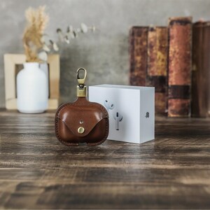 May include: A brown leather AirPod case with a brass clasp and button closure. The case is next to a white box with a graphic of white AirPods. The background includes a vase, books, and a wooden surface.