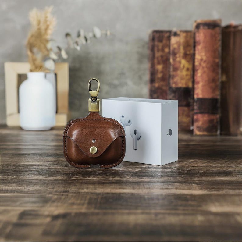 May include: A brown leather AirPod case with a brass clasp and button closure. The case is next to a white box with a graphic of white AirPods. The background includes a vase, books, and a wooden surface.