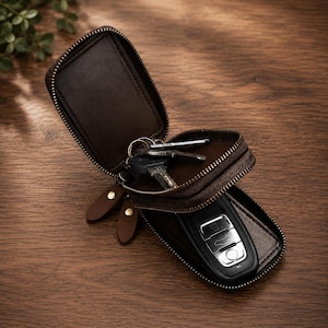 Könnte beinhalten: Ein dunkelbraunes Schlüsseletui aus Leder mit Reißverschluss, das eine offene Klappe und ein Hauptfach mit einem schwarzen Autoschlüssel mit silbernen Tasten und einem Metallring enthält. Das Etui hat zwei braune Leder-Reißverschlussanhänger.