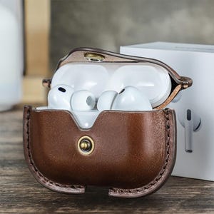 May include: A brown leather case with white earbuds inside. The case is open, revealing the earbuds, and has a snap closure. A white box is in the background, and a single earbud is visible to the right.