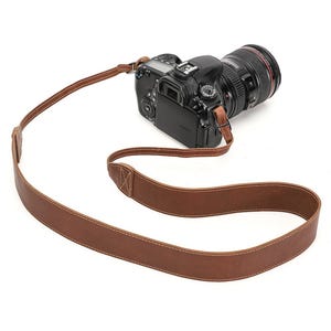 May include: A brown leather camera strap with white stitching is attached to a black DSLR camera with a large lens. The strap is made of a single piece of leather and has a simple, classic design.
