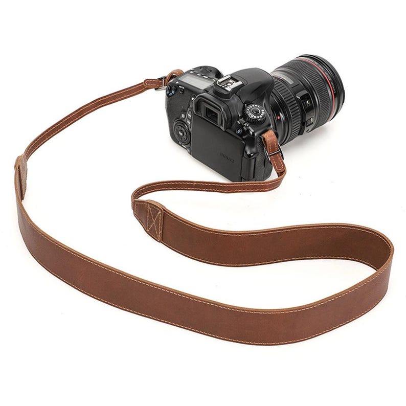 May include: A brown leather camera strap with white stitching is attached to a black DSLR camera with a large lens. The strap is made of a single piece of leather and has a simple, classic design.
