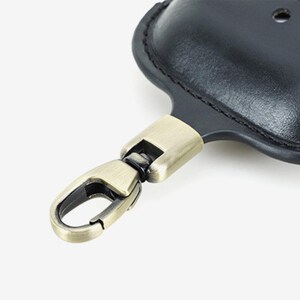 May include: Black leather key holder with a metal clasp. The key holder has a rounded shape with a stitched border. The metal clasp is a bronze color and is attached to the key holder with a swivel mechanism.