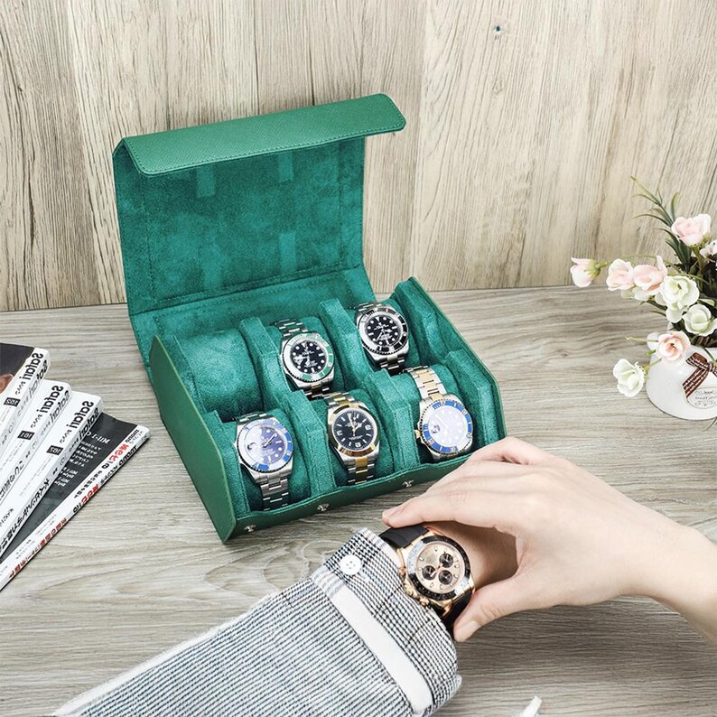 May include: A green leather watch case with six compartments, each holding a wristwatch. The watches have silver, gold, and black bands and faces. The case is open, revealing a plush green interior. A person is wearing a gold watch.
