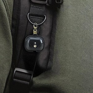 May include: Black leather AirPod case with a snap closure, attached to a black backpack strap with a metal clasp. The case has two small, round details on the front.