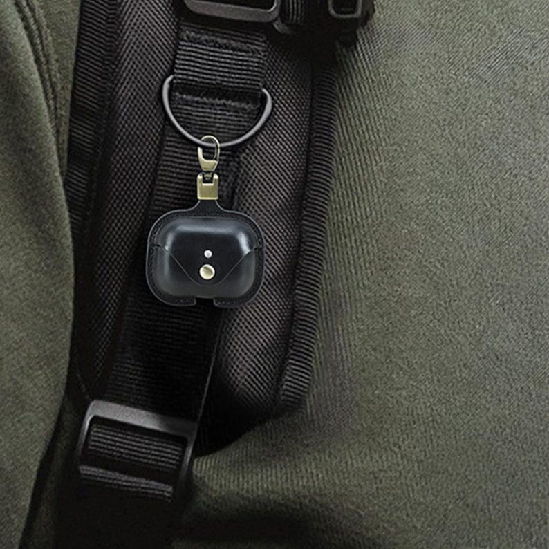 May include: Black leather AirPod case with a snap closure, attached to a black backpack strap with a metal clasp. The case has two small, round details on the front.