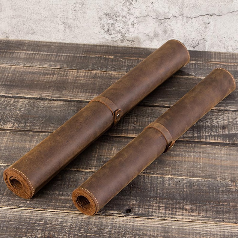 May include: Two rolled-up brown leather mats with leather straps and buckle closures. The mats are lying on a wooden surface. The leather has a distressed look.
