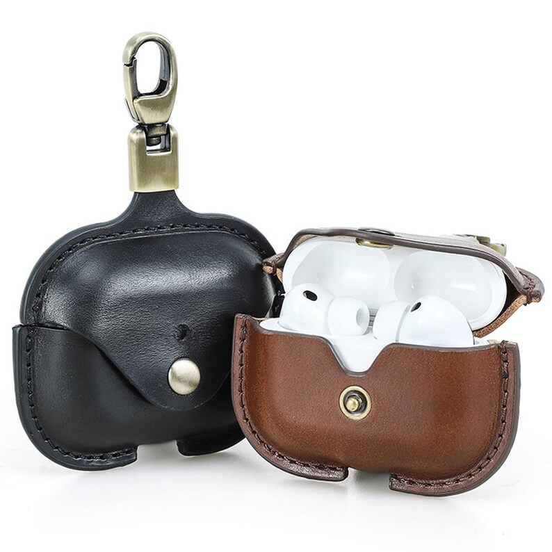 May include: Two leather AirPod cases. One is black with a silver clasp and a silver button closure. The other is brown, open, and contains white earbuds. Both cases have a metal clasp for attaching to a keychain.