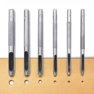 Könnte beinhalten: Ein Set aus sechs silberfarbenen Lochstanzen aus Metall, die in einer Reihe angeordnet sind, jeweils mit einem strukturierten, gerändelten Griff und einer hohlen zylindrischen Spitze, über einem beigen Lederstreifen mit sechs unterschiedlich großen Löchern.