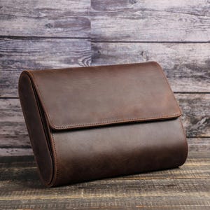 May include: A brown leather travel case with a flap closure. The case is rectangular with rounded edges and is made of smooth, dark brown leather. The case is set against a wooden background.