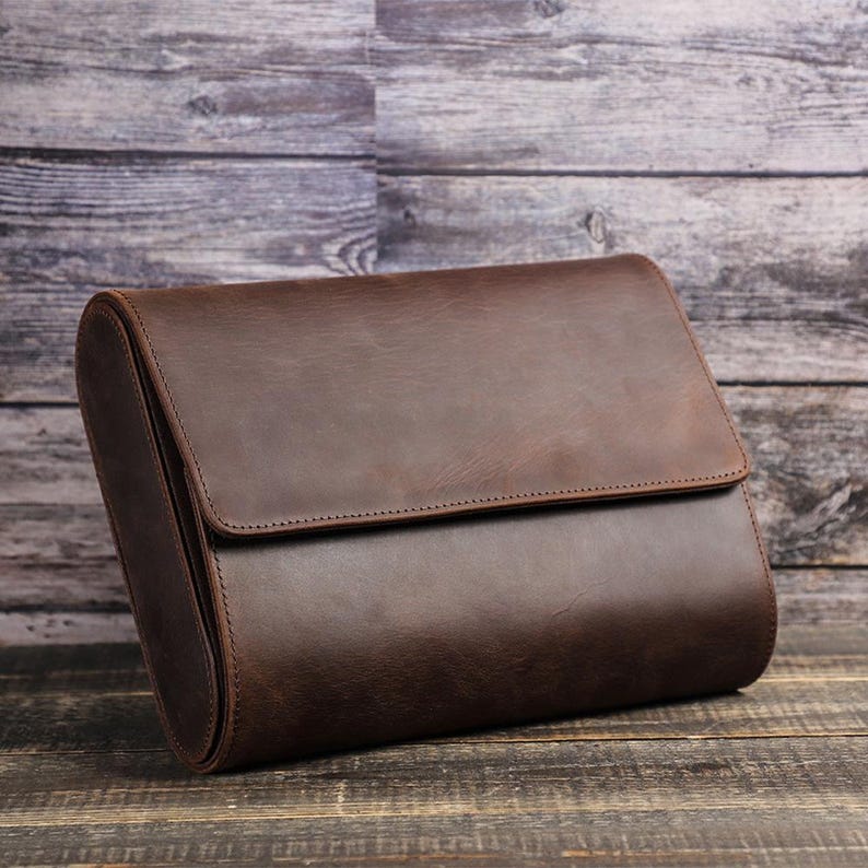 May include: A brown leather travel case with a flap closure. The case is rectangular with rounded edges and is made of smooth, dark brown leather. The case is set against a wooden background.