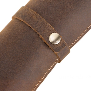 May include: Close-up of a brown leather case with a strap and metal snap closure. The leather has a distressed appearance with visible grain and texture. The stitching is visible along the edges.