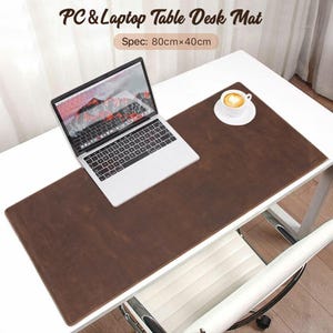 May include: A brown PC and laptop table desk mat, 80cm x 40cm, with a laptop and a cup of coffee on a white desk. The mat is rectangular and made of a textured material. The laptop is open, displaying a mountain landscape.