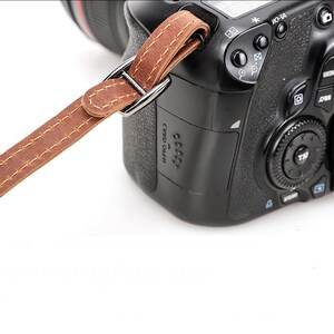 May include: A brown leather camera strap attached to a black DSLR camera. The strap is stitched and connected to the camera with a silver metal ring. The camera has various buttons and dials.