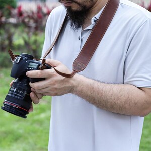 May include: A brown leather camera strap with a padded shoulder section and stitched detailing. The strap is attached to a black DSLR camera with a lens. The photographer is wearing a light gray polo shirt.