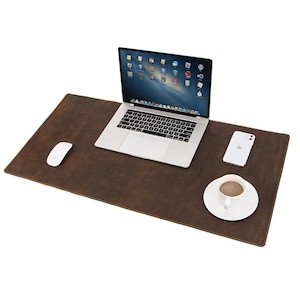 May include: A brown leather desk mat with a laptop, white mouse, smartphone, and a cup of coffee on a white saucer. The laptop is open, displaying a screen with icons. The desk mat is rectangular.