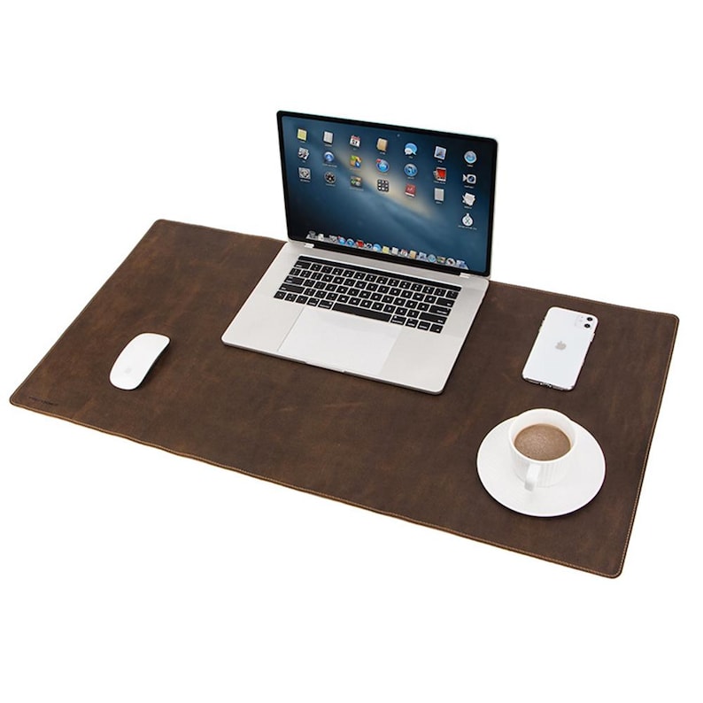May include: A brown leather desk mat with a laptop, white mouse, smartphone, and a cup of coffee on a white saucer. The laptop is open, displaying a screen with icons. The desk mat is rectangular.