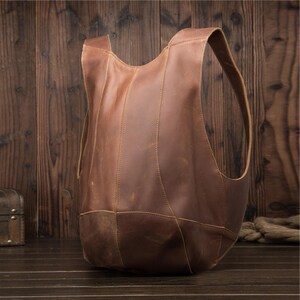 May include: A brown leather backpack with a unique, open design. The bag features a rounded base and a single, wide shoulder strap. The leather has a distressed appearance, with visible stitching and variations in color. The bag is designed to be worn over one shoulder.