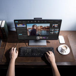 May include: A computer monitor displaying video editing software, with a keyboard and mouse on a brown desk. A white smartphone and a cup of coffee are also on the desk. The monitor shows a woman's portrait and video editing tools.