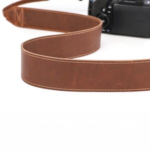 May include: A brown leather camera strap with cream-colored stitching. The strap is made of a thick, durable leather and is designed to attach to a camera. The strap is shown against a white background.