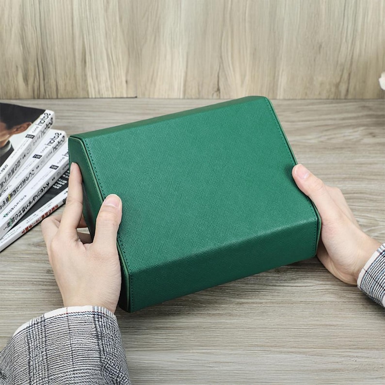 May include: A rectangular, emerald green box with a textured surface. The box is held by two hands, and the background includes a wooden surface and a stack of magazines.