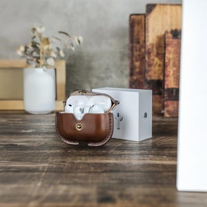 May include: A brown leather case with a gold button clasp, holding white wireless earbuds. A white box with a logo is next to the case. The case is on a wooden surface.