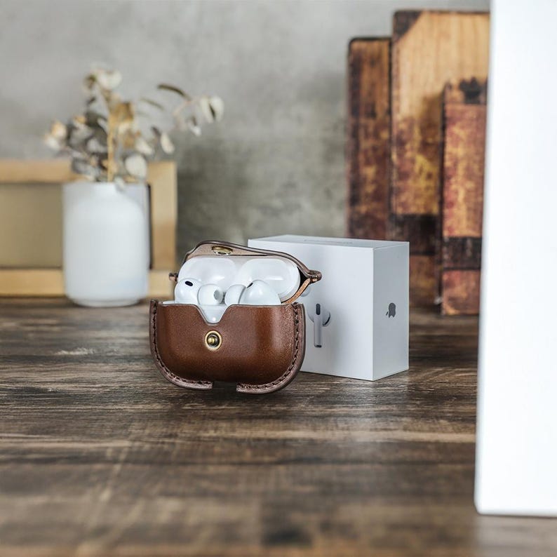 May include: A brown leather case with a gold button clasp, holding white wireless earbuds. A white box with a logo is next to the case. The case is on a wooden surface.