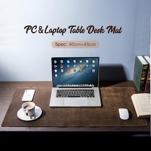 May include: A brown leather desk mat with the text "PC & Laptop Table Desk Mat" and "Spec: 90cm x 45cm". A laptop, a white mouse, a smartphone, and a coffee cup on a saucer are on the mat.
