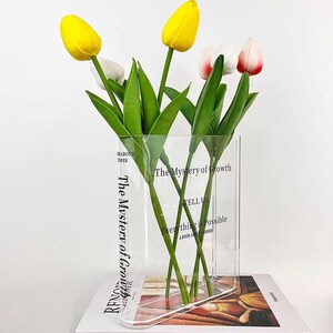 Könnte beinhalten: Eine transparente, rechteckige Vase mit gebogenem Boden, die gelbe und weiße Tulpen mit grünen Blättern enthält. Die Vase trägt den Text "The Mystery of Growth" und "Everything is Possible". Die Vase steht auf einem Buch.