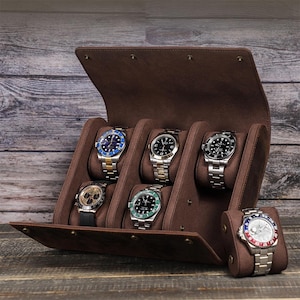 Puede incluir: Un estuche de cuero marrón para relojes con seis relojes. Los relojes tienen correas plateadas y doradas, y esferas negras, azules y verdes. El estuche está abierto, mostrando los relojes sobre cojines marrones.