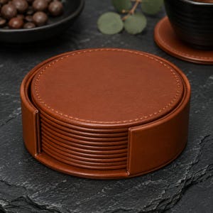Peut inclure: Une pile de dessous de verre ronds en similicuir marron, rangés dans un support circulaire assorti. Chaque dessous de verre présente une surface lisse de couleur unie avec des coutures visibles sur le bord extérieur, créant un ensemble uniforme.