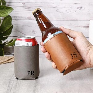 Pode incluir: Dois isoladores de bebidas de couro personalizados, um segurando uma garrafa de cerveja e o outro uma lata de refrigerante. O isolador marrom tem as iniciais "JS" e o isolador cinza tem "PR". A garrafa de cerveja é marrom e a lata de refrigerante é vermelha.