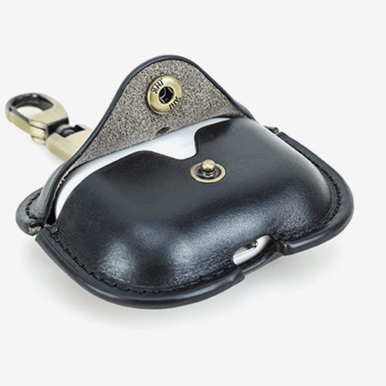 May include: Black leather AirPod case with a snap closure and a metal clasp. The case is open, revealing the white interior. The case has a stitched border and a vintage brass-colored clasp.